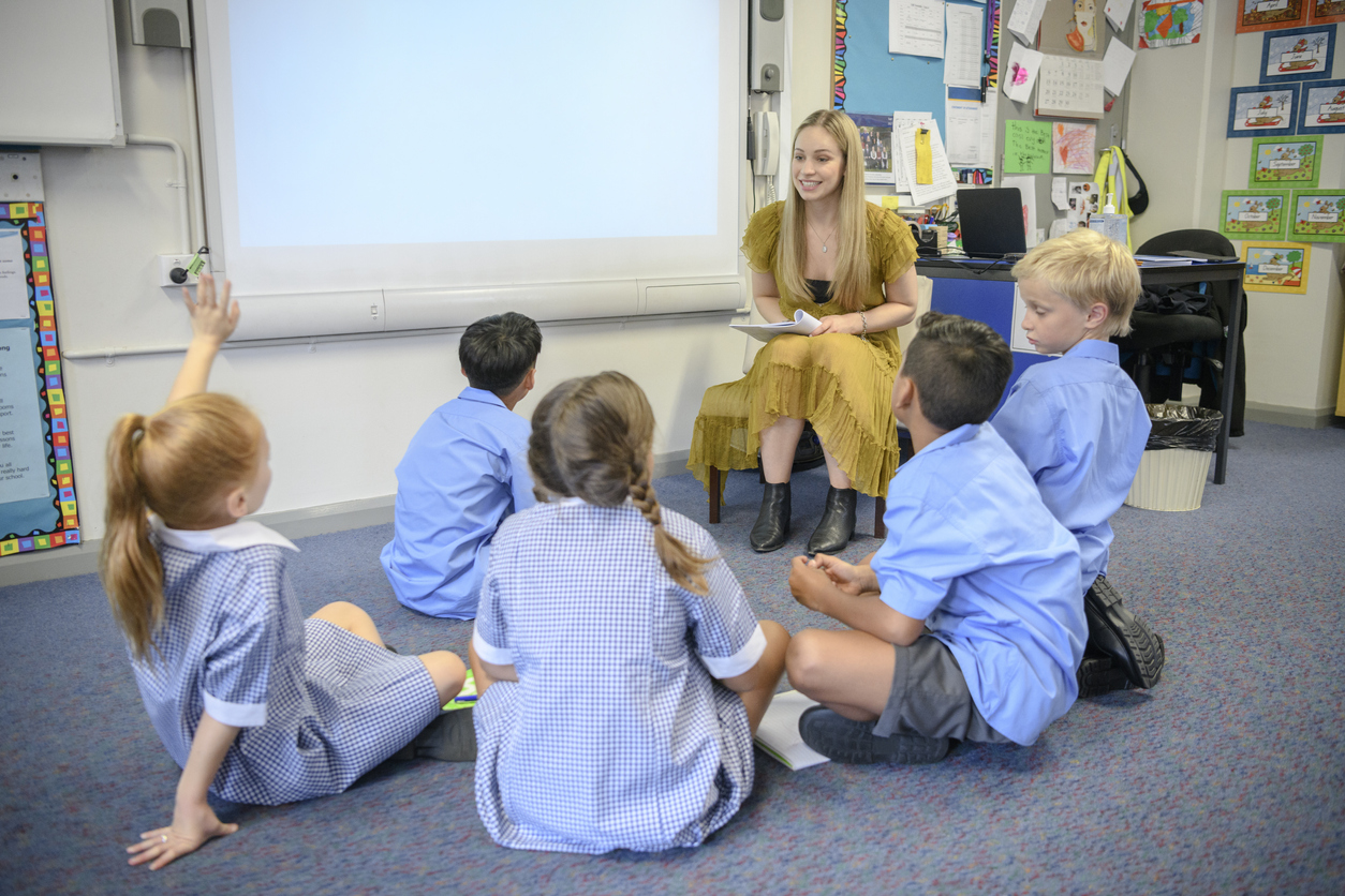 Young woman teaching multi racial students in primary school on the carpet